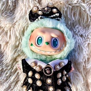May include: A whimsical doll with a mint green furry hood and a black dress adorned with pearls and rhinestones. The doll has large, expressive eyes and a mischievous grin, and wears a black crown with pearl accents.
