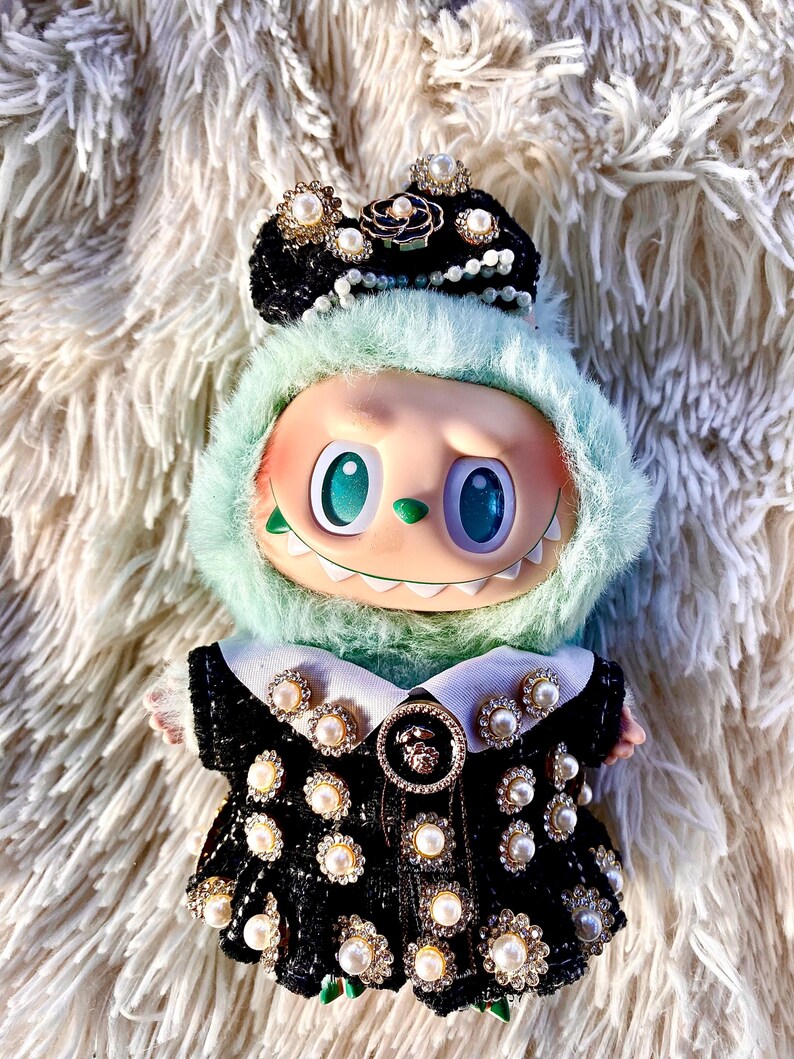 May include: A whimsical doll with a mint green furry hood and a black dress adorned with pearls and rhinestones. The doll has large, expressive eyes and a mischievous grin, and wears a black crown with pearl accents.