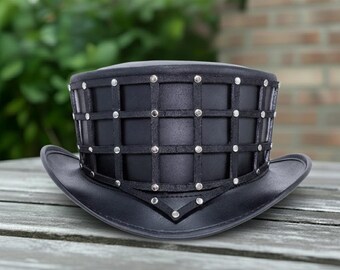 May include: A black top hat with a wide brim, featuring a grid-like design on the crown. The hat is adorned with silver-colored studs at the intersections of the grid. The hat is made of a dark material and sits on a wooden surface.