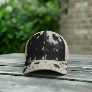 May include: A baseball cap with a black and white cowhide pattern on the crown and brim. The cap has a neutral-colored front panel and a curved brim. The hat is designed for casual wear.