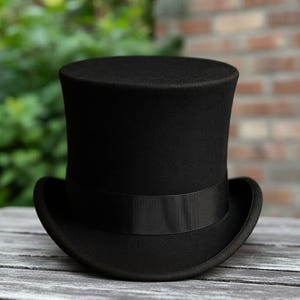 May include: A traditional black top hat, featuring a broad brim and a black band. The hat is crafted from a felt-like material and rests on a weathered wooden surface. The background includes foliage and a brick wall.