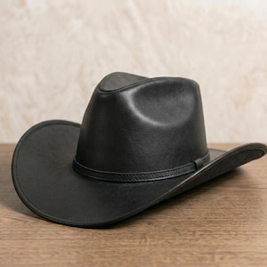 May include: A black leather cowboy hat featuring a pinched crown and a curved brim. The hat includes a thin, matching black leather band encircling the base of the crown, providing a clean and uniform appearance across the entire structure.