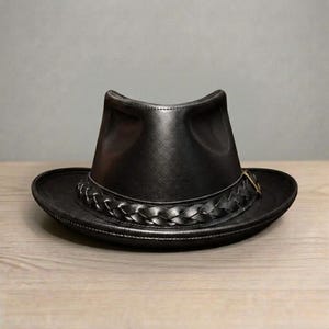 Peut inclure: Chapeau en cuir noir avec un large bord et une bande tressée. Le chapeau a une forme de fedora classique avec une couronne légèrement creusée. Le chapeau est posé sur une surface en bois.