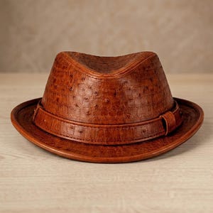 Può includere: Un cappello fedora in pelle marrone con finitura strutturata. Il cappello presenta una corona strutturata, un'ampia tesa e una fascia intorno alla base. Un accessorio classico.