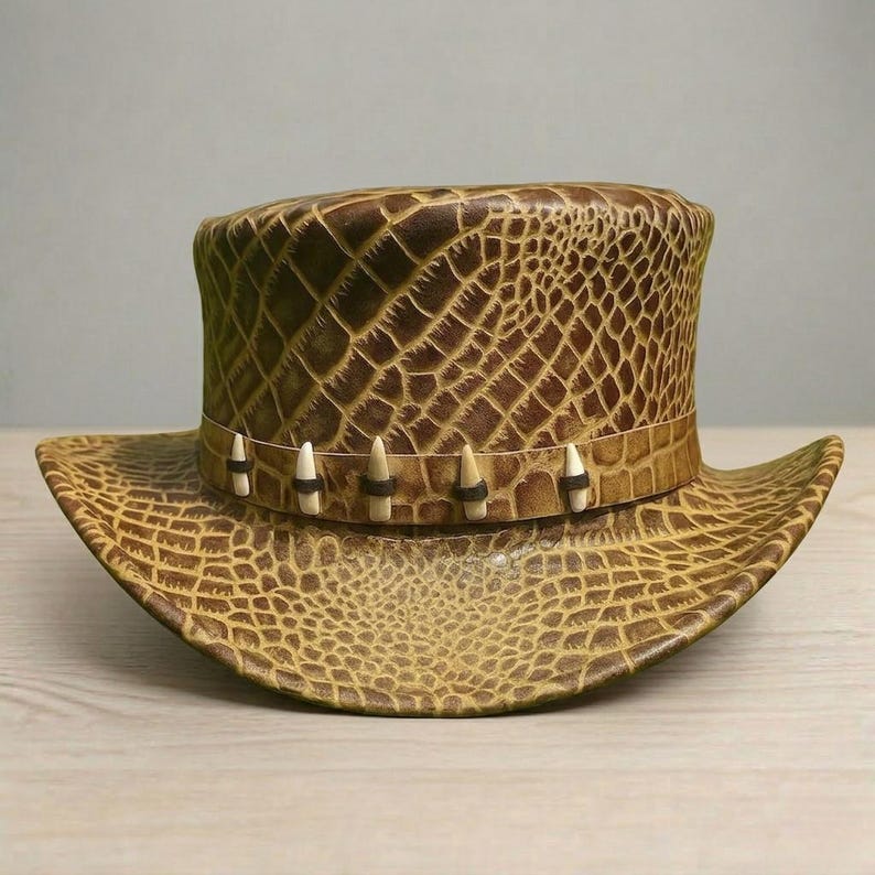 May include: A brown, textured hat with a wide brim and a tall, cylindrical crown. The hat features a band with decorative elements and a pattern that resembles crocodile skin. The hat is a light brown color.