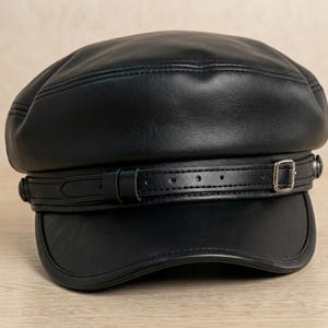 May include: A black cap with a smooth finish, featuring a structured crown and a curved brim. A decorative black strap with a silver-toned metal buckle and matching side studs wraps horizontally around the base of the crown above the brim.