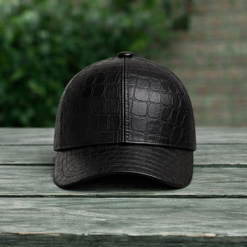 K&ouml;nnte beinhalten: Schwarze Baseballkappe mit einem strukturierten Muster in Krokodilleder-Optik. Die Kappe hat einen gebogenen Schirm und einen Knopf oben. Sie besteht aus leder&auml;hnlichem Material und liegt auf einer Holzoberfl&auml;che.