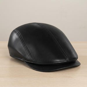 Könnte beinhalten: Eine schwarze Leder-Flatcap mit gebogenem Schirm. Die Kappe hat eine glatte, glänzende Oberfläche und ein klassisches, stromlinienförmiges Design. Die Kappe liegt auf einer Holzoberfläche.