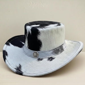 Puede incluir: Sombrero de vaquero con estampado de piel de vaca en blanco y negro. El sombrero tiene una copa redondeada y un ala ancha. Una banda plateada con un bot&oacute;n decorativo rodea la base de la copa. El sombrero est&aacute; hecho de un material suave y texturizado.