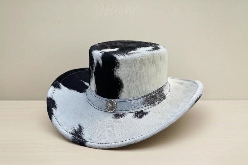 Puede incluir: Sombrero de vaquero con estampado de piel de vaca en blanco y negro. El sombrero tiene una copa redondeada y un ala ancha. Una banda plateada con un bot&oacute;n decorativo rodea la base de la copa. El sombrero est&aacute; hecho de un material suave y texturizado.