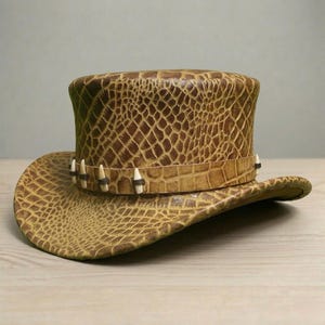 May include: A brown leather top hat with a wide brim, featuring a textured, crocodile-like pattern. The hatband is adorned with small, white, tooth-like embellishments. The hat's design evokes a rustic, adventurous aesthetic.