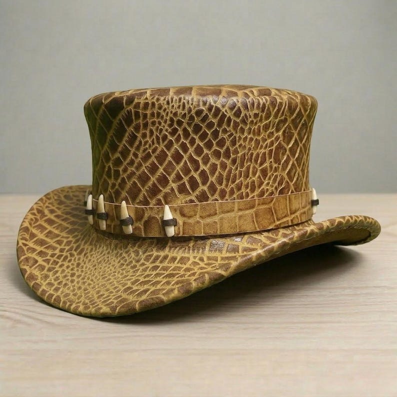 May include: A brown leather top hat with a wide brim, featuring a textured, crocodile-like pattern. The hatband is adorned with small, white, tooth-like embellishments. The hat's design evokes a rustic, adventurous aesthetic.