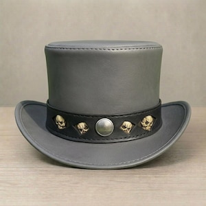 Puede incluir: Un sombrero de copa gris con una banda negra adornada con motivos de calaveras doradas. El sombrero tiene un ala ancha y un detalle de botón plateado en la banda. El sombrero está hecho de un material liso similar al cuero.