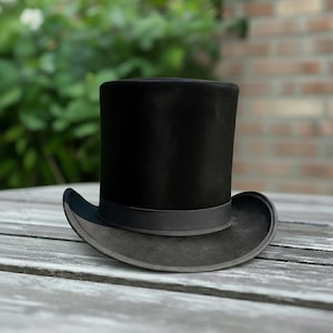 Könnte beinhalten: Ein schwarzer Zylinderhut mit breiter Krempe und dunkelgrauem Band. Der Hut ist aus einem filzartigen Material gefertigt und liegt auf einer Holzoberfläche. Ein klassisches formelles Accessoire.