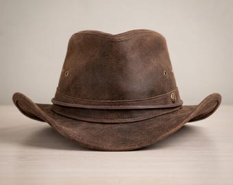 May include: A brown leather hat with a wide brim and a slightly indented crown. The hat features a leather band around the base of the crown and small metal eyelets. A small decorative emblem is on the side of the hat.