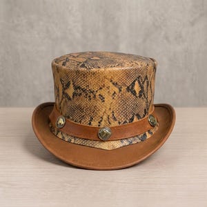 Puede incluir: Un sombrero de copa de cuero marrón con un estampado de piel de serpiente en la copa. El sombrero tiene un ala de cuero marrón y una banda con botones metálicos decorativos. Un accesorio de moda único.