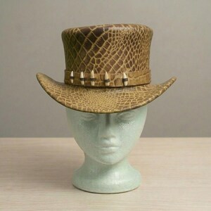 May include: A brown top hat with a textured, reptile-like pattern. The hat features a wide brim and a band adorned with decorative teeth-like accents. The hat is displayed on a light green mannequin head, set against a neutral background.