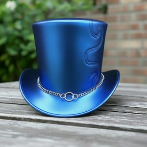 May include: A glossy blue top hat with a silver chain around the brim. The hat features a stitched, curved design on the side. It rests on a wooden surface.