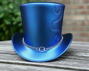 May include: A shiny blue top hat with a silver chain around the brim. The hat has a curved design stitched on the side. The hat is sitting on a wooden surface.