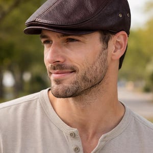 Könnte beinhalten: Braune Leder-Newsboy-Cap mit runder Krone und kleinem, steifem Schirm. Die Kappe hat eine strukturierte Oberfläche und ist mit kleinen Metallnieten an der Seite befestigt. Ein klassischer Stil.