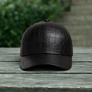 K&ouml;nnte beinhalten: Schwarze Baseballkappe mit einem strukturierten Muster in Krokodilleder-Optik. Die Kappe hat einen gebogenen Schirm und einen Knopf oben. Sie besteht aus leder&auml;hnlichem Material und liegt auf einer Holzoberfl&auml;che.