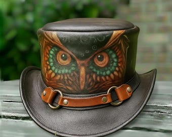Puede incluir: Un sombrero de copa de cuero marrón oscuro con ala ancha. El sombrero presenta un diseño detallado de cara de búho en tonos marrón, naranja y verde. Una correa de cuero marrón con anillos y remaches metálicos rodea el sombrero.