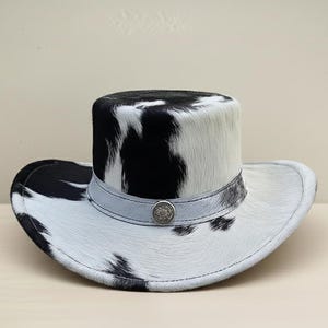 Puede incluir: Sombrero de vaquero con un patr&oacute;n de piel de vaca en blanco y negro. El sombrero presenta una corona cil&iacute;ndrica y un ala ancha. Una banda gris claro con un bot&oacute;n decorativo plateado rodea la corona. El sombrero est&aacute; dise&ntilde;ado para la protecci&oacute;n solar y el estilo.