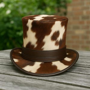 May include: A top hat with a cowhide pattern in shades of brown and cream. The hat features a brown leather band and brim, creating a classic and rustic look. The hat is likely for costume or fashion purposes.
