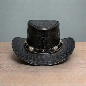 May include: A black cowboy hat with a textured, crocodile-like pattern. The hat has a decorative band with silver-coloured studs around the crown. The hat is on a wooden surface.