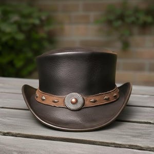 Puede incluir: Un sombrero de copa de cuero negro con ala y banda marrones. La banda está adornada con tachuelas plateadas y un medallón decorativo plateado y cobrizo. El sombrero se exhibe sobre una superficie de madera envejecida.