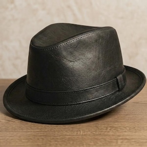 Puede incluir: Un sombrero fedora negro confeccionado con un material liso y texturizado. El sombrero presenta una copa estructurada con un pliegue central, un ala ancha y una banda negra a juego que rodea la base de la copa con costuras precisas.
