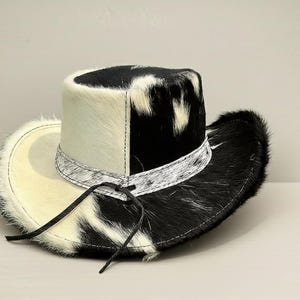 Puede incluir: Un sombrero de vaquero con un patr&oacute;n &uacute;nico de piel de vaca en blanco y negro. El sombrero presenta una corona y un ala de color crema, con una banda estampada en blanco y negro alrededor de la base de la corona. Tiene una correa de barbilla de cuero.