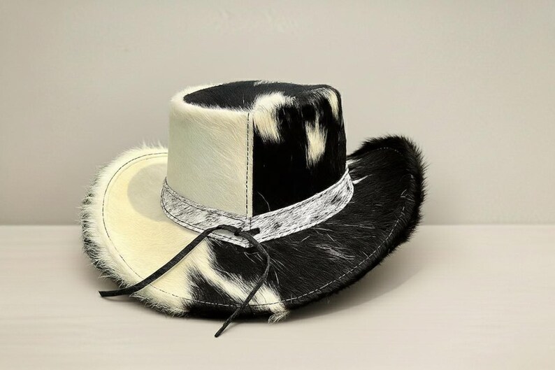 Puede incluir: Un sombrero de vaquero con un patr&oacute;n &uacute;nico de piel de vaca en blanco y negro. El sombrero presenta una corona y un ala de color crema, con una banda estampada en blanco y negro alrededor de la base de la corona. Tiene una correa de barbilla de cuero.