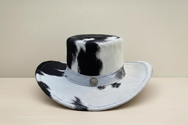Puede incluir: Sombrero de vaquero con un patr&oacute;n de piel de vaca en blanco y negro. El sombrero tiene una copa alta y redondeada y un ala ancha. Una banda gris claro con un bot&oacute;n decorativo plateado rodea la copa.