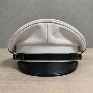 Pode incluir: Um chapéu de couro branco com aba e faixa pretas. O chapéu tem uma copa arredondada e uma aba plana e estruturada. A faixa é de couro preto e envolve a base do chapéu. O chapéu está sobre uma superfície de madeira clara.