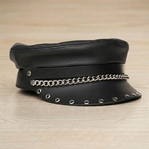 May include: Black leather hat with a flat top and a short, curved brim. A silver chain is wrapped around the hat, just above the brim. The hat has silver eyelets around the brim.