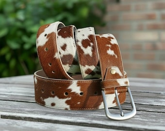 May include: A brown and white cowhide belt with a silver buckle. The belt is made of genuine leather and features a natural cowhide pattern. The belt is shown coiled on a wooden surface.