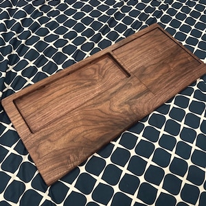 May include: A rectangular wooden serving tray with a dark brown finish. The tray features two recessed sections, one long and rectangular, and another smaller, square-shaped. The wood grain is visible throughout the tray.
