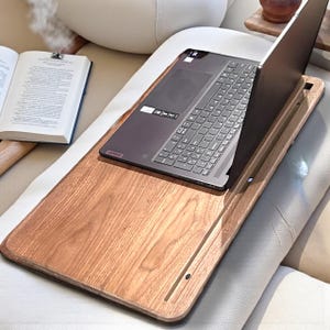May include: A wooden laptop desk with a laptop on top. The desk is rectangular with a raised edge to prevent items from sliding off. The laptop is dark gray and open, displaying the keyboard and screen. An open book is visible in the background.