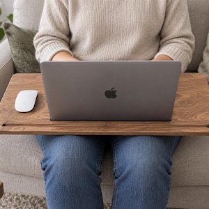 May include: A rectangular wooden lap desk with a dark brown wood grain finish, featuring a horizontal slot for holding devices, supporting a silver laptop with an Apple logo and a white computer mouse on its flat surface.