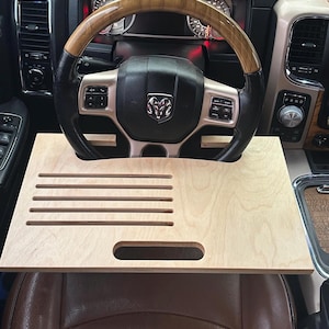 Puede incluir: Un escritorio de madera clara para volante de coche con forma rectangular. El escritorio tiene cuatro ranuras paralelas y un recorte para el asa. El escritorio está colocado sobre un asiento de coche de cuero marrón, con el volante al fondo.