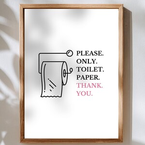 Op de afbeelding: Een ingelijste print met een minimalistisch ontwerp. Het kunstwerk toont een zwarte lijntekening van een toiletpapierrol en de tekst "PLEASE. ONLY. TOILET. PAPER. THANK. YOU." in zwart en roze. Het frame is van hout.