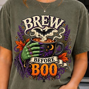 May include: A dark olive green t-shirt featuring a graphic print of a green witch hand holding a black mug with steam and a bat silhouette. The text reads BREW BEFORE BOO in white and orange lettering, surrounded by purple lavender and orange leaves.