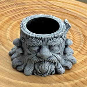 May include: A gray, 3D-printed planter shaped like a tree stump with a face carved into it. The face has a long beard, closed eyes, and mushroom accents. The planter sits on a wooden surface.
