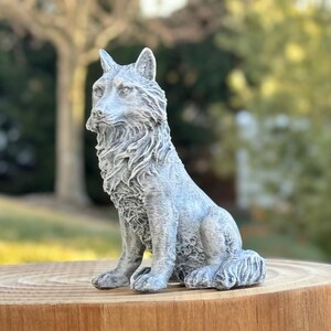 May include: A detailed, gray wolf statue in a seated position. The sculpture has a textured surface, capturing the fur and features of the animal. It is placed on a wooden surface, with a blurred outdoor background.