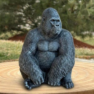 May include: A detailed, gray gorilla statue in a seated position. The sculpture has a textured surface, with visible muscle definition on the chest and arms. The gorilla's face shows a serious expression. The statue is placed on a wooden surface, with a blurred green background.
