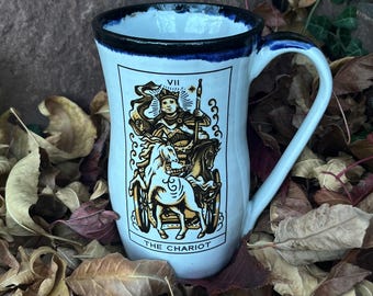 Op de afbeelding: Een witte keramische mok met een zwarte en blauwe rand, met een tarotkaartontwerp van "The Chariot". De mok heeft een gebogen handvat en is omgeven door gevallen bladeren.
