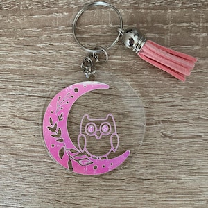 May include: A clear acrylic keyring with a pink crescent moon design, floral details, and an owl silhouette. It has a silver keyring, chain, and a pink tassel.