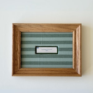 May include: A rectangular wooden frame with a light brown wood grain finish containing a fabric mat with horizontal sage green and light green stripes. A small white fortune cookie slip with the text "Great thoughts come from the heart" is centered.