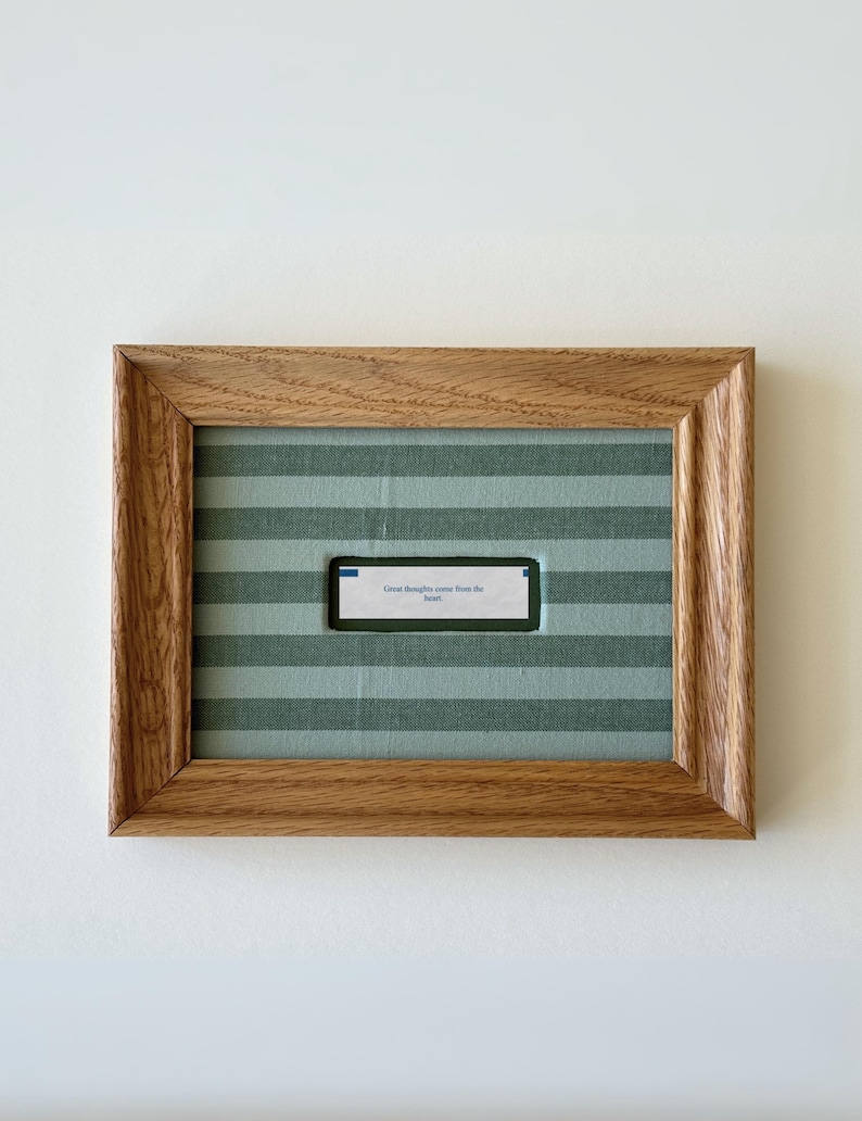 May include: A rectangular wooden frame with a light brown wood grain finish containing a fabric mat with horizontal sage green and light green stripes. A small white fortune cookie slip with the text "Great thoughts come from the heart" is centered.